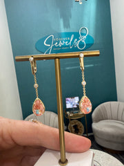A pair of dangling earrings displayed on a gold stand, held by a hand in a jewelry studio. The earrings feature gold-toned metal with clear stones and pink teardrop-shaped earrings 